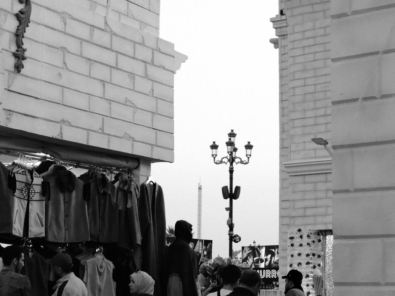 A bustling street market scene with clothing displays and people walking, framed by stone architecture.
