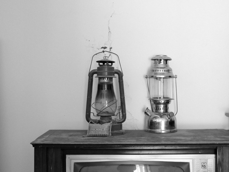 Two vintage lanterns on a wooden surface against a plain wall, one is glass and metal, the other is metal.