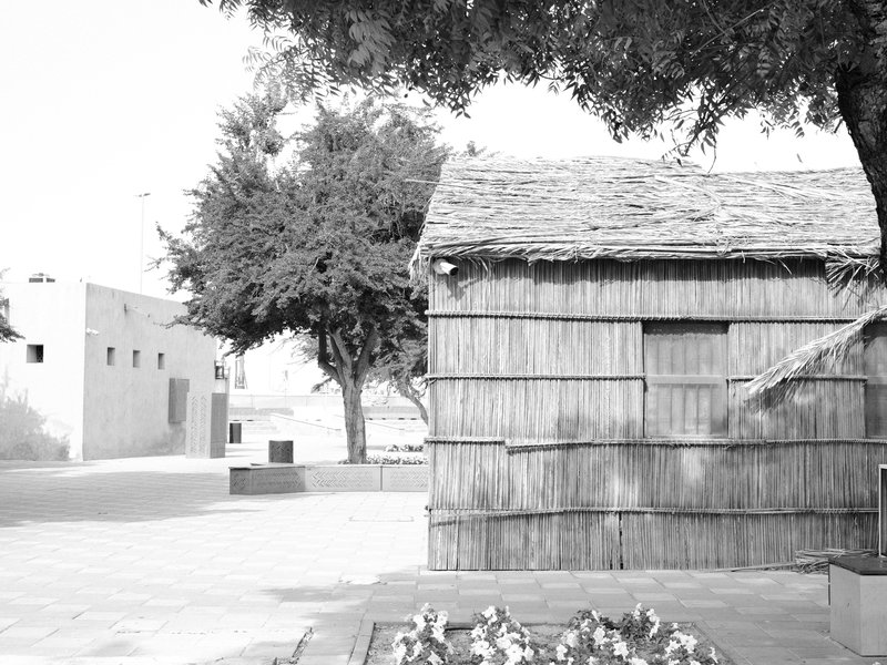 Black and white photo of a rustic wooden structure beside a tree, with a distant building in a quiet setting.