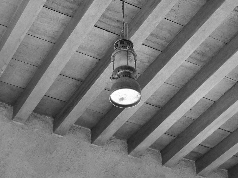 A vintage lantern hanging from a wooden ceiling with exposed beams, in black and white.