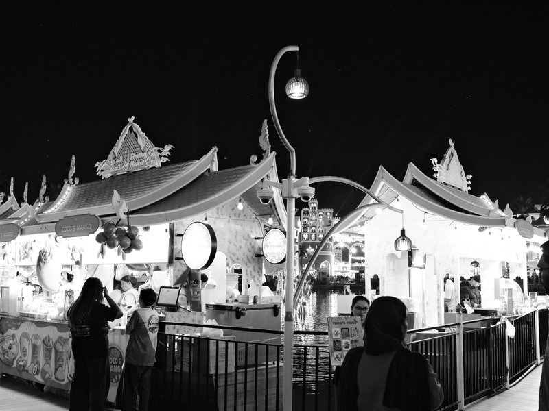 Night scene of illuminated stalls at a fair, with people interacting and festive decorations.