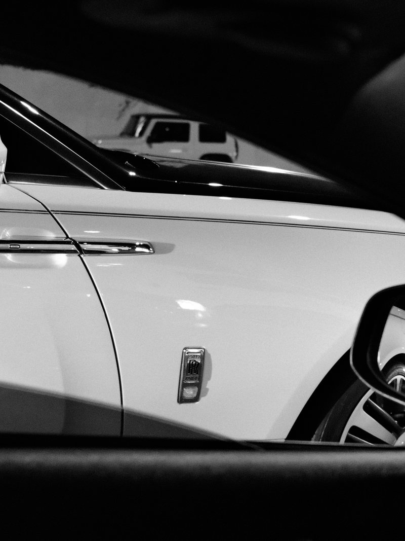 Close-up of a white luxury car's side, showcasing its sleek design and emblem, with reflections in the window.