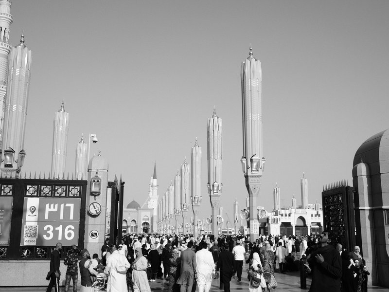 Crowd of people in a bustling outdoor market with tall decorative structures and clear blue sky.