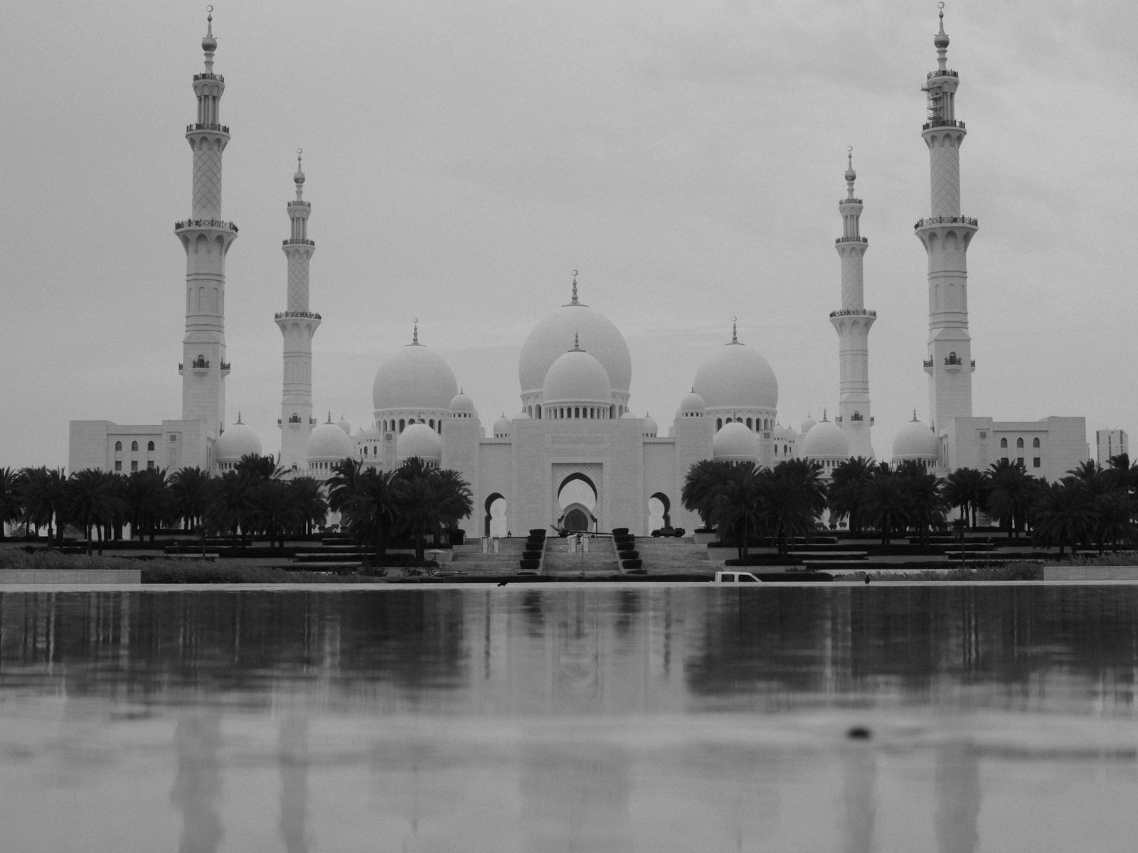 A grand mosque with multiple domes and minarets reflected in a calm water body under a cloudy sky.
