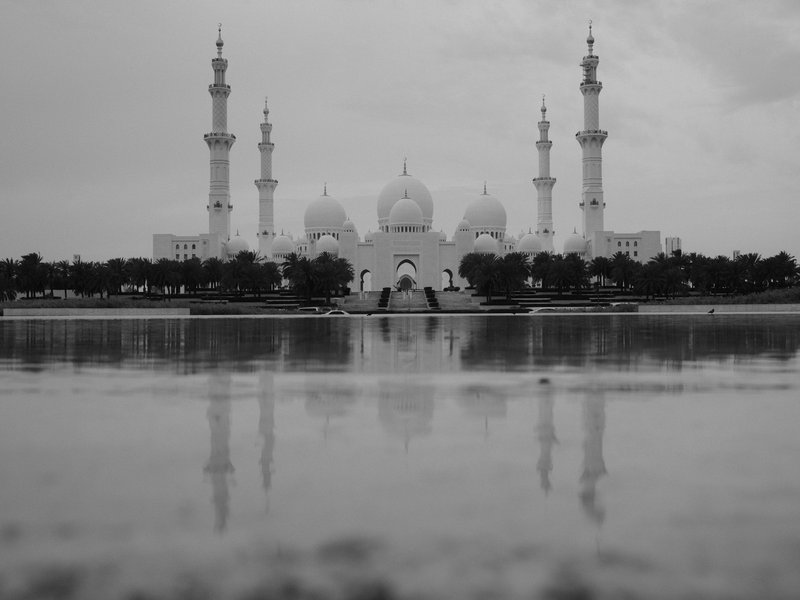 A grand mosque with multiple minarets reflected in a calm water surface under a cloudy sky.