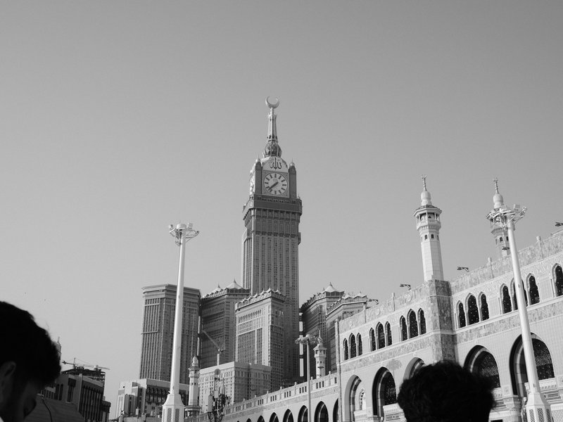 A tall clock tower rises above a historic building in a clear sky, with people in the foreground.