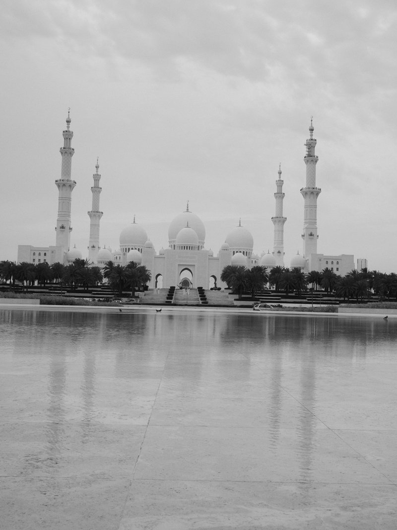 A grand white mosque with multiple minarets reflected in a calm water surface under a cloudy sky.