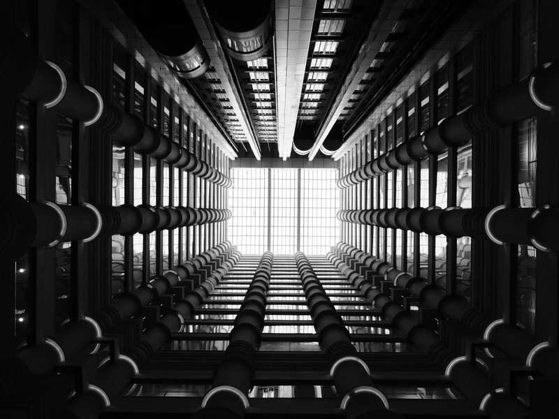 Looking up through a modern architectural structure with pipes and beams, illuminated by a bright skylight.