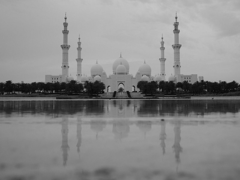 Black and white image of a grand mosque with multiple minarets and a reflective pool in the foreground.