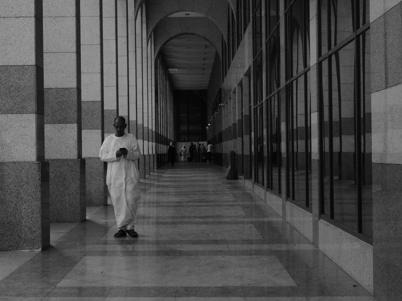 A person in white clothing stands in a long corridor with reflective glass walls and a tiled floor.