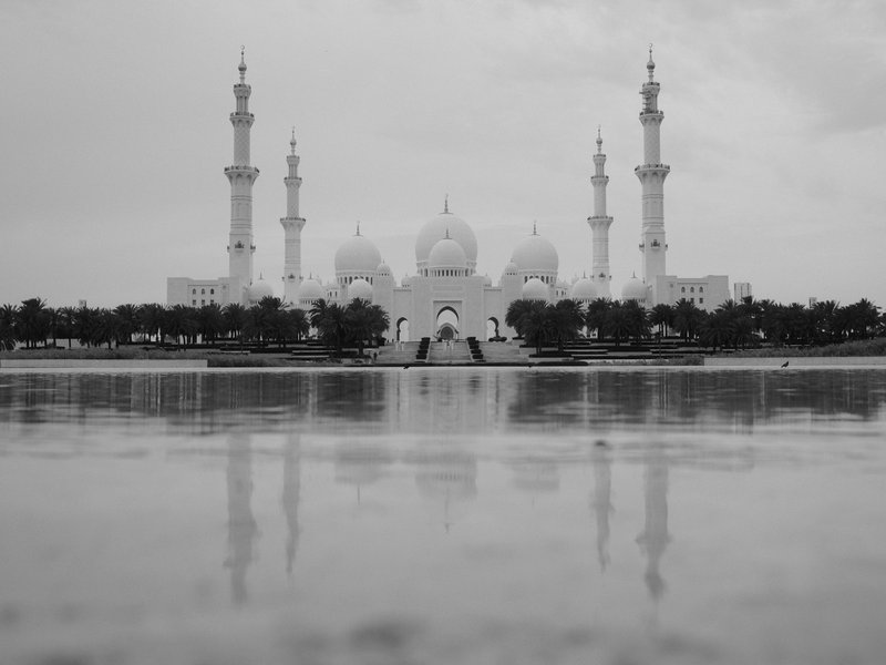 A grand mosque with multiple minarets reflected in a wet surface under a cloudy sky.