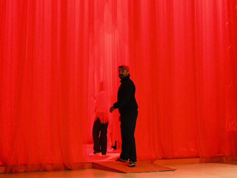 Two figures silhouetted against a vibrant red curtain in a dimly lit space.