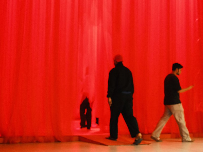 Two figures walking through a translucent red curtain in a dimly lit space.