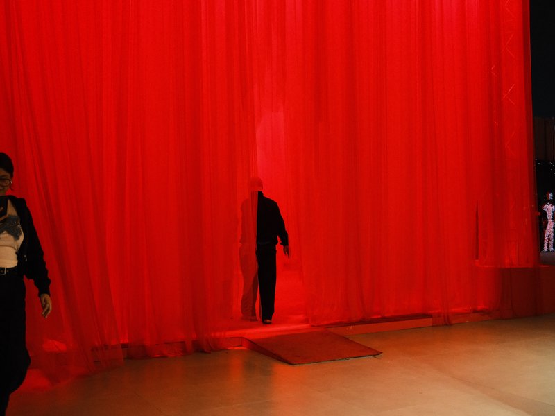 A figure emerges from behind a bright red curtain in a dimly lit space.