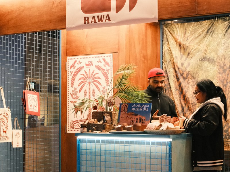 A vendor and a customer interact at a small shop with a wooden counter and decorative posters.