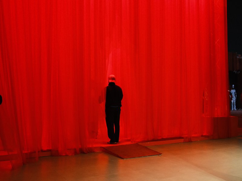 A person standing in front of a sheer red curtain, partially illuminated, creating a dramatic atmosphere.
