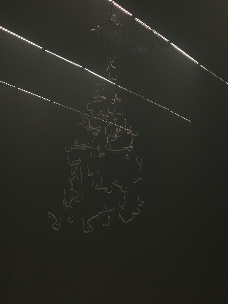 A dark scene featuring illuminated Arabic script hanging from the ceiling, with faint light beams above.