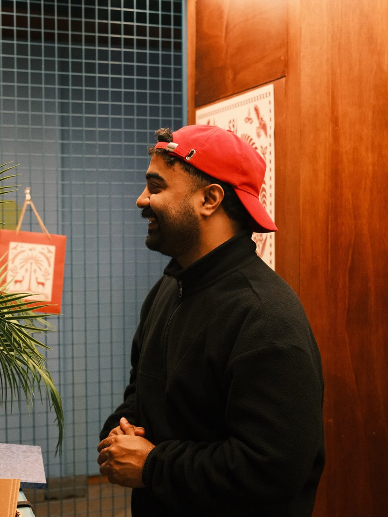 A man in a red cap stands beside a wooden wall, with a plant and framed art in the background.