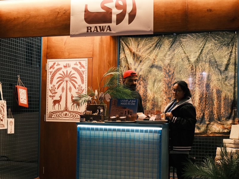 Two people at a market stall with a decorative backdrop and a sign reading 'RAWA'.