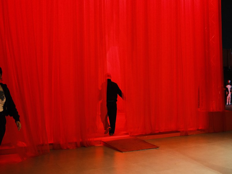 A figure walking through a red curtain in a dimly lit space, with another figure visible in the background.