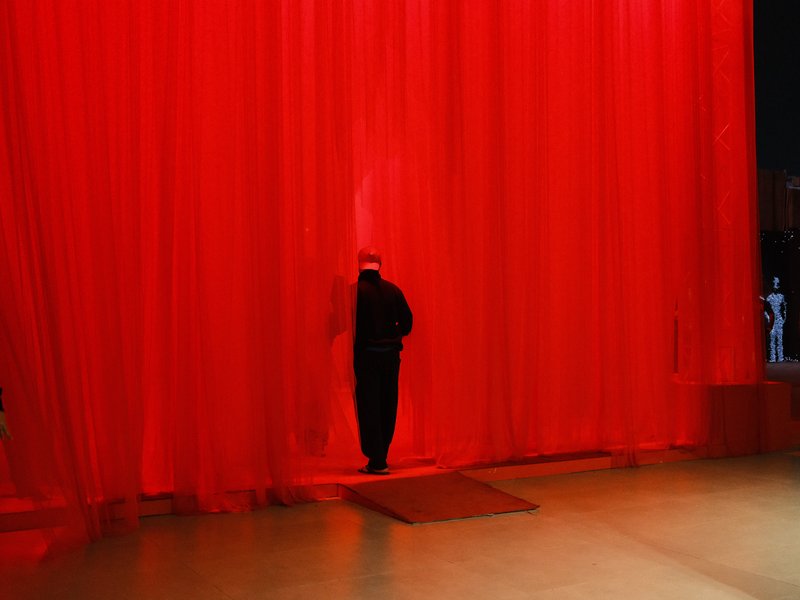 A figure in black steps through a vibrant red curtain, creating a dramatic contrast in the scene.
