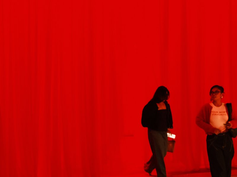 Two figures walking in front of a vibrant red backdrop, one looking down at a device.