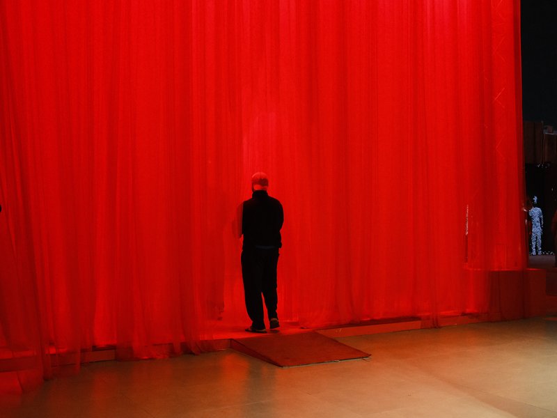 A figure in black stands before a vibrant red curtain, creating a dramatic and moody atmosphere.