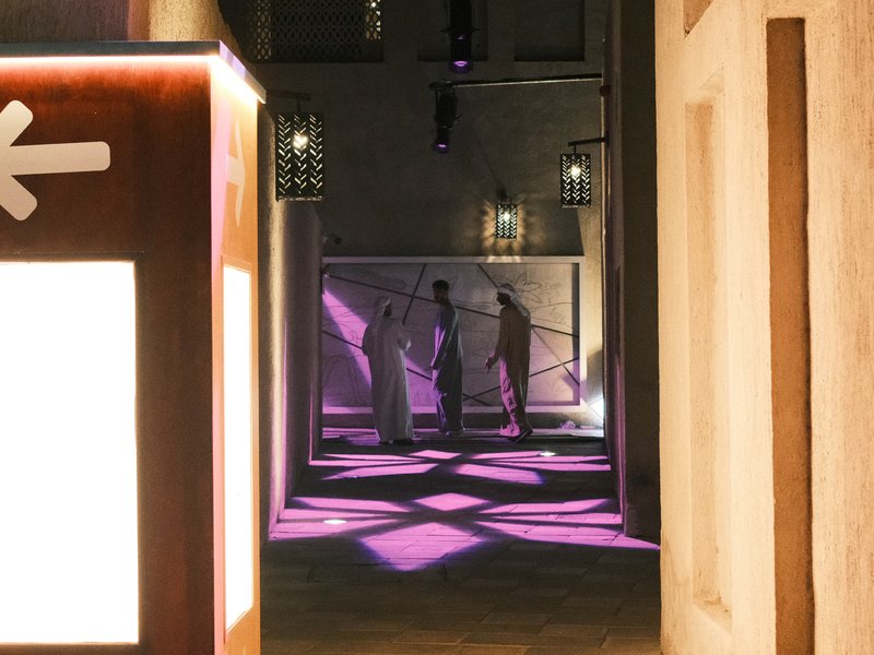 Silhouetted figures in traditional attire walking through a narrow alley illuminated by purple light.
