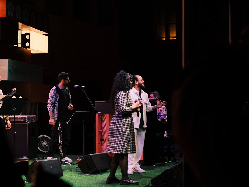 A live music performance with singers and musicians on stage, illuminated by stage lights.
