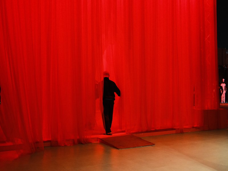 A figure in black steps through a red curtain, creating a dramatic scene with vibrant colors.