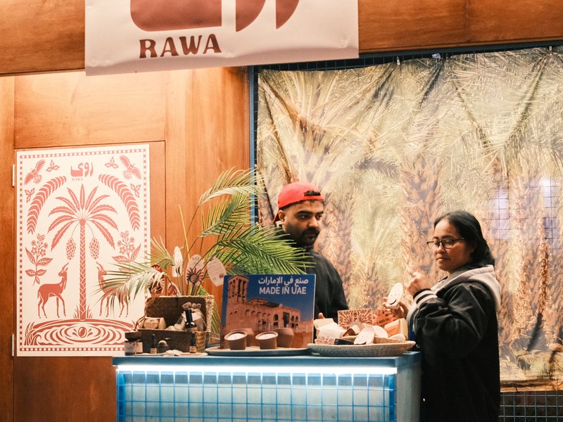 Two people at a food stall with a sign reading 'Rawa', surrounded by decorative elements.