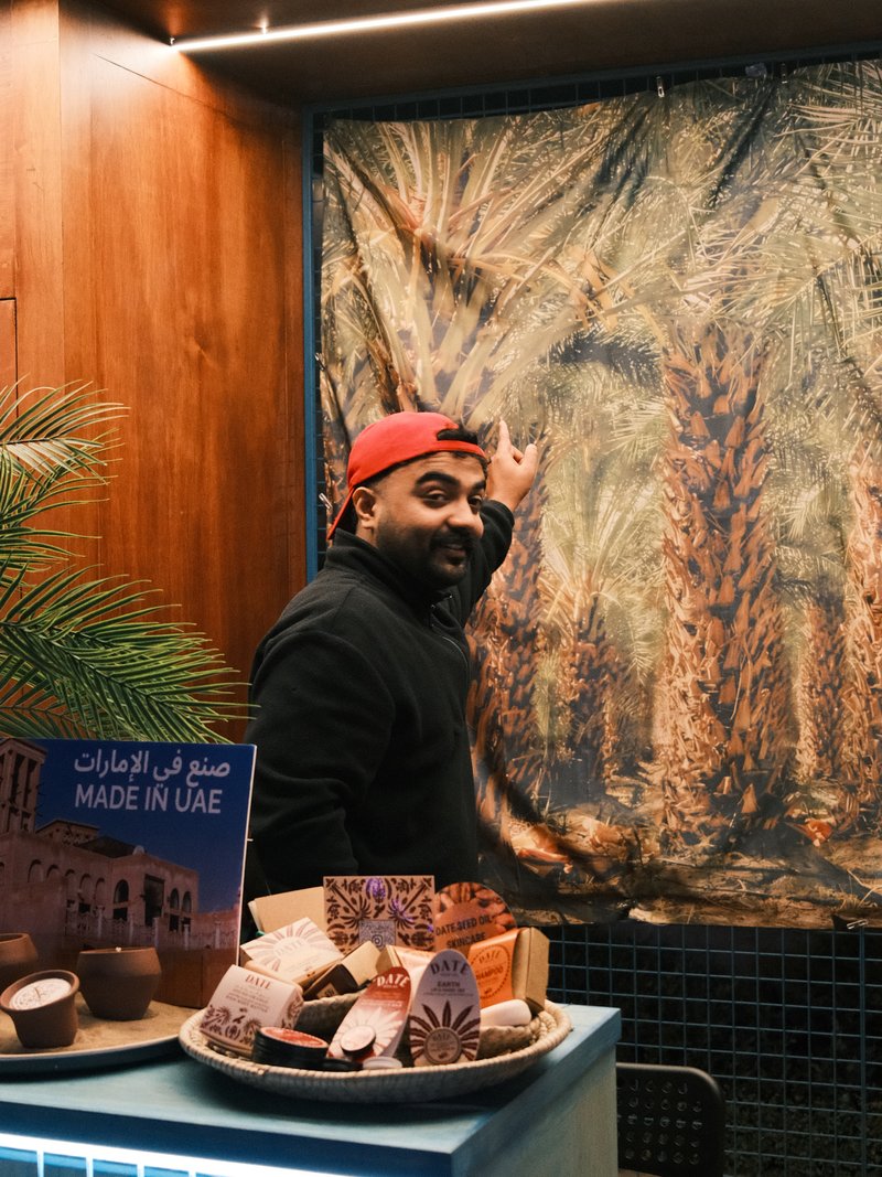 A man in a red cap gestures towards a palm tree backdrop in a cozy indoor setting.