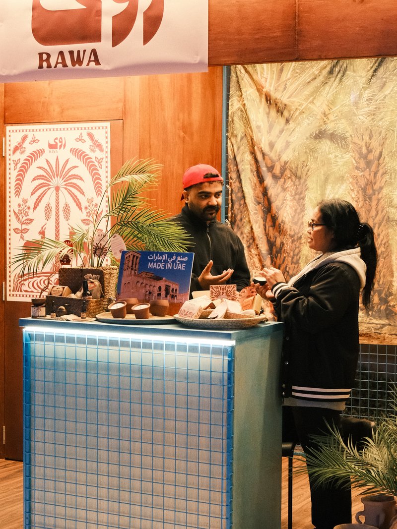 A vendor and a customer engage in conversation at a market stall decorated with plants and cultural motifs.