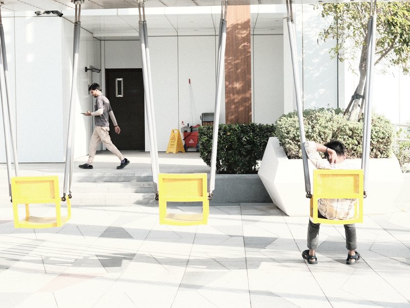 Two swings in a modern outdoor setting with a person walking by and another sitting on a swing.
