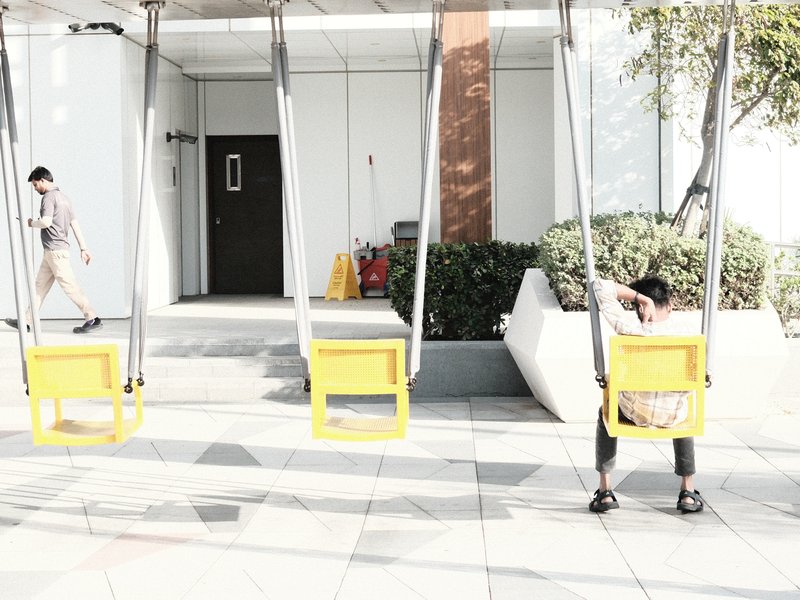 Two yellow swings in an urban setting with a person walking in the background and another sitting on a swing.