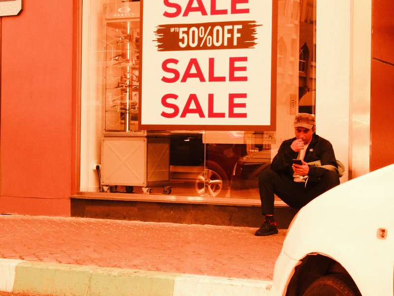 A man sitting outside a store with a large sale sign, wearing sunglasses and holding a drink.