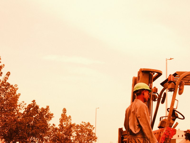 A construction worker stands beside a tractor under a warm, orange sky with trees in the foreground.