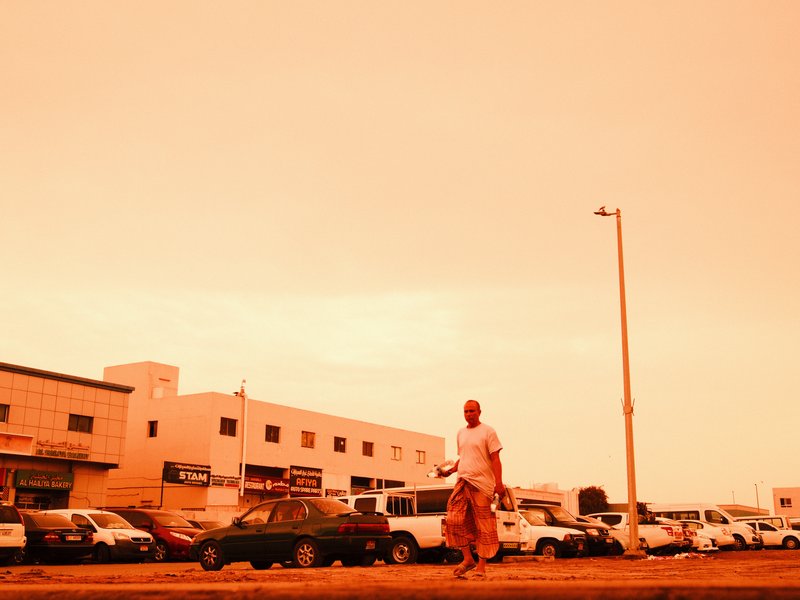 A man walking on a street with buildings and parked cars under an orange sky.