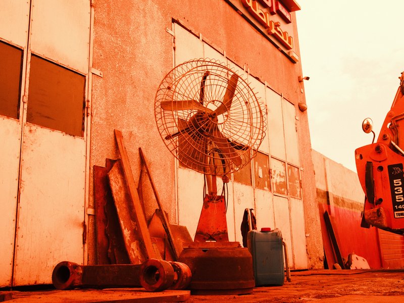 An industrial fan stands against a weathered wall, surrounded by wooden planks and a blue container.