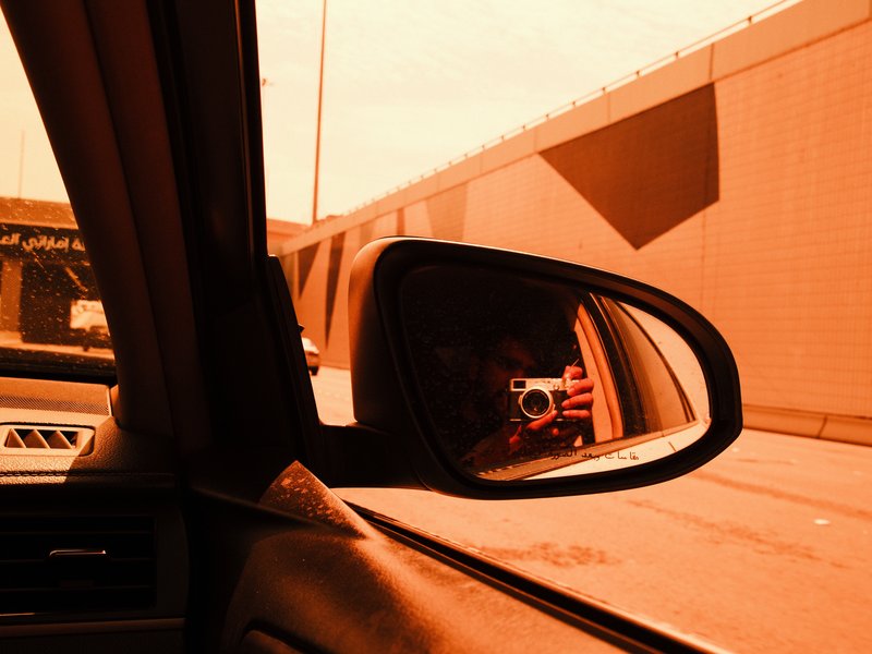 A car's side mirror reflecting an orange-toned urban scene with a wall and a hint of a vehicle.
