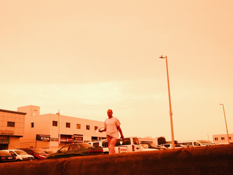 A person walking on a street with a warm orange sky and buildings in the background.