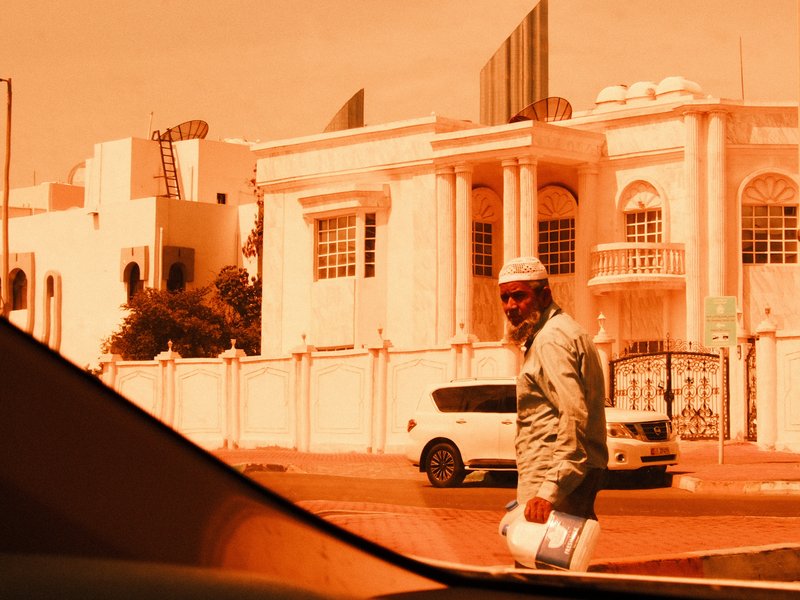 A man in traditional attire walking on a street with a white building in the background under an orange hue.