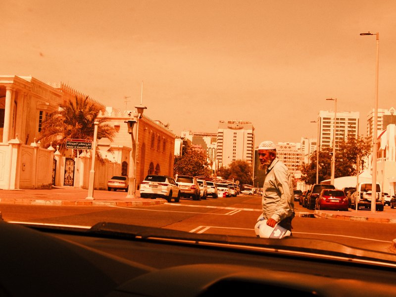A street scene with a person walking across the road, urban buildings in the background under an orange hue.