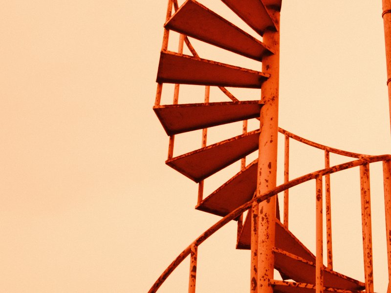 A close-up of a spiral staircase with an orange tint against a pale sky.