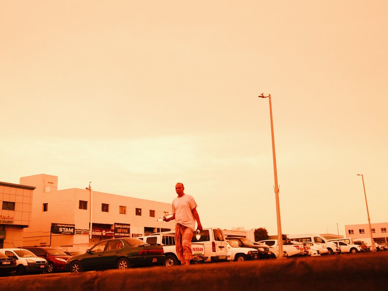 A person walking along a street with buildings and parked cars under an orange sky.
