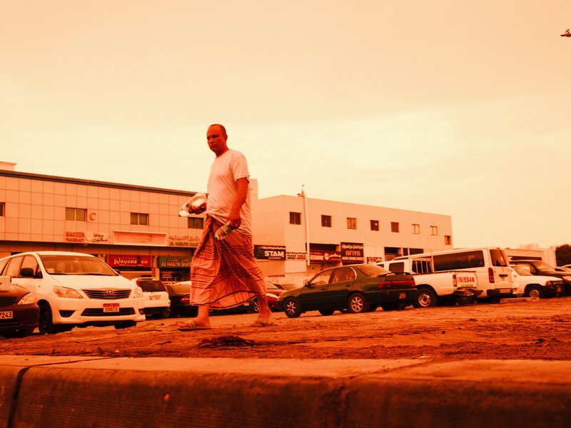 A man walking on a street with vehicles parked nearby under an orange-tinted sky.