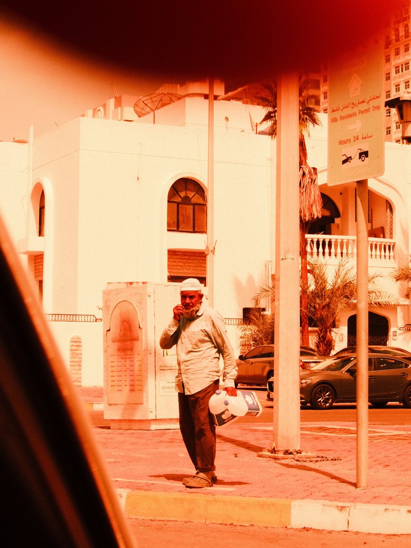 A man in a light shirt and hat stands on a street, holding a phone, with buildings and palm trees in the background.