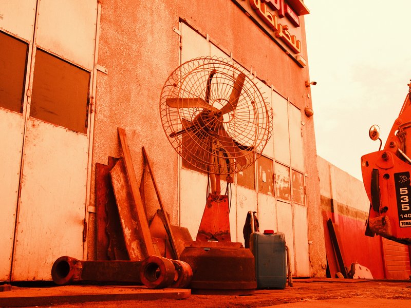 An industrial fan stands against a weathered wall with wooden planks and a blue container nearby.