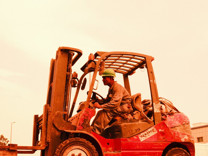 A worker operating a red forklift in an industrial setting under an orange sky.