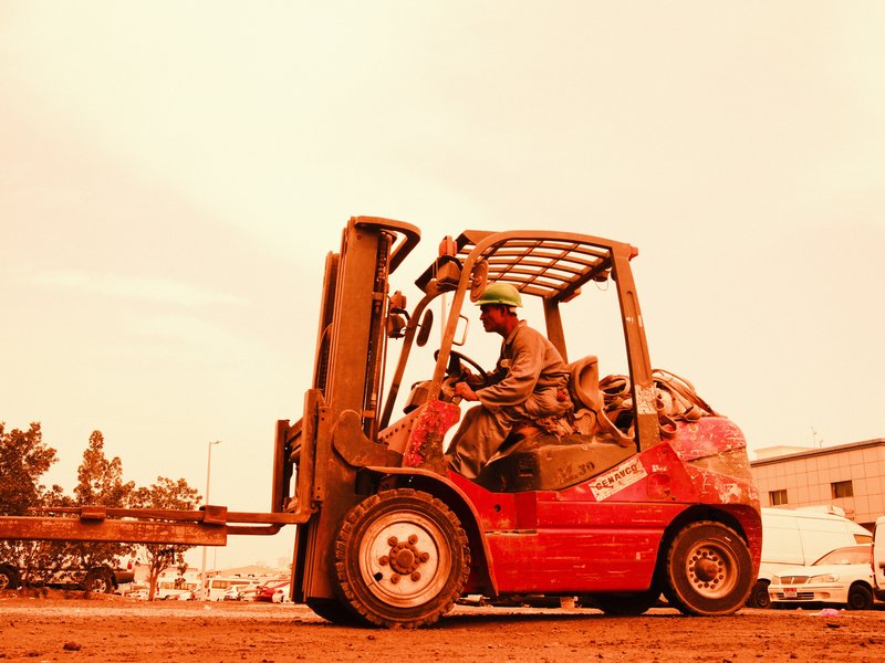 A red forklift operated by a worker in a dusty outdoor setting with an orange hue.